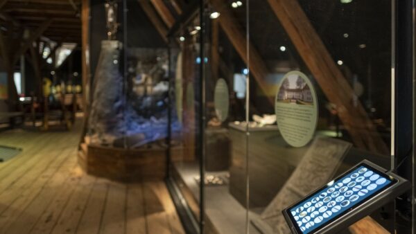 Bilde Av En Rekke Museumsgjenstander Beskyttet Bak En Glassvegg. Det Henger Et Nettbrett På Siden Av Glassveggen. På Nettbrettet Vises Små Bilder Av Hver Gjenstand. Det Er Mulig å Trykke På Bildene For å Få Informasjon Om Hver Enkelt Gjenstand Bak Glassmonteret. Jeg Er Nødt Til å Finne En Alternativ Formidlingsløsning For Blinde Og Svaksynte.