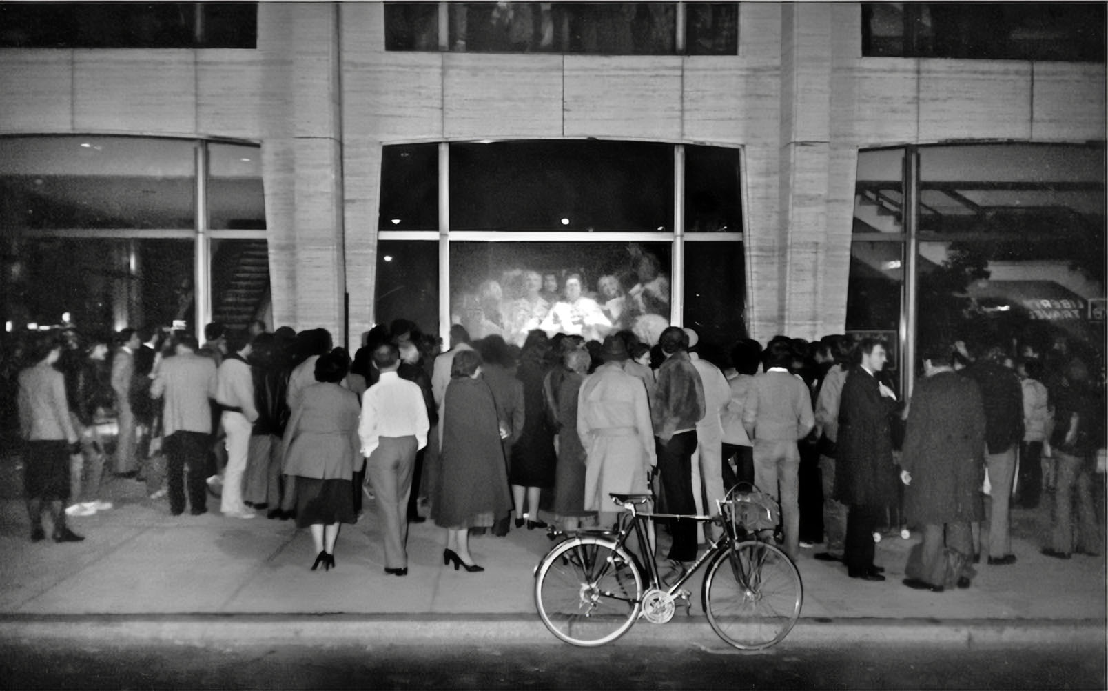 A black and white photograph depicts a large crowd of individuals gathered outside a building during nighttime, engrossed in a large illuminated window display. Inside the window, a projection shows multiple faces, a representation of the groundbreaking "Hole-in-Space" art installation. The onlookers, display a sense of wonder and curiosity. A solitary bicycle is parked in the foreground, adding depth to the scene. The image captures a pivotal moment in media art history, reflecting the intersection of technology, art, and human connection.