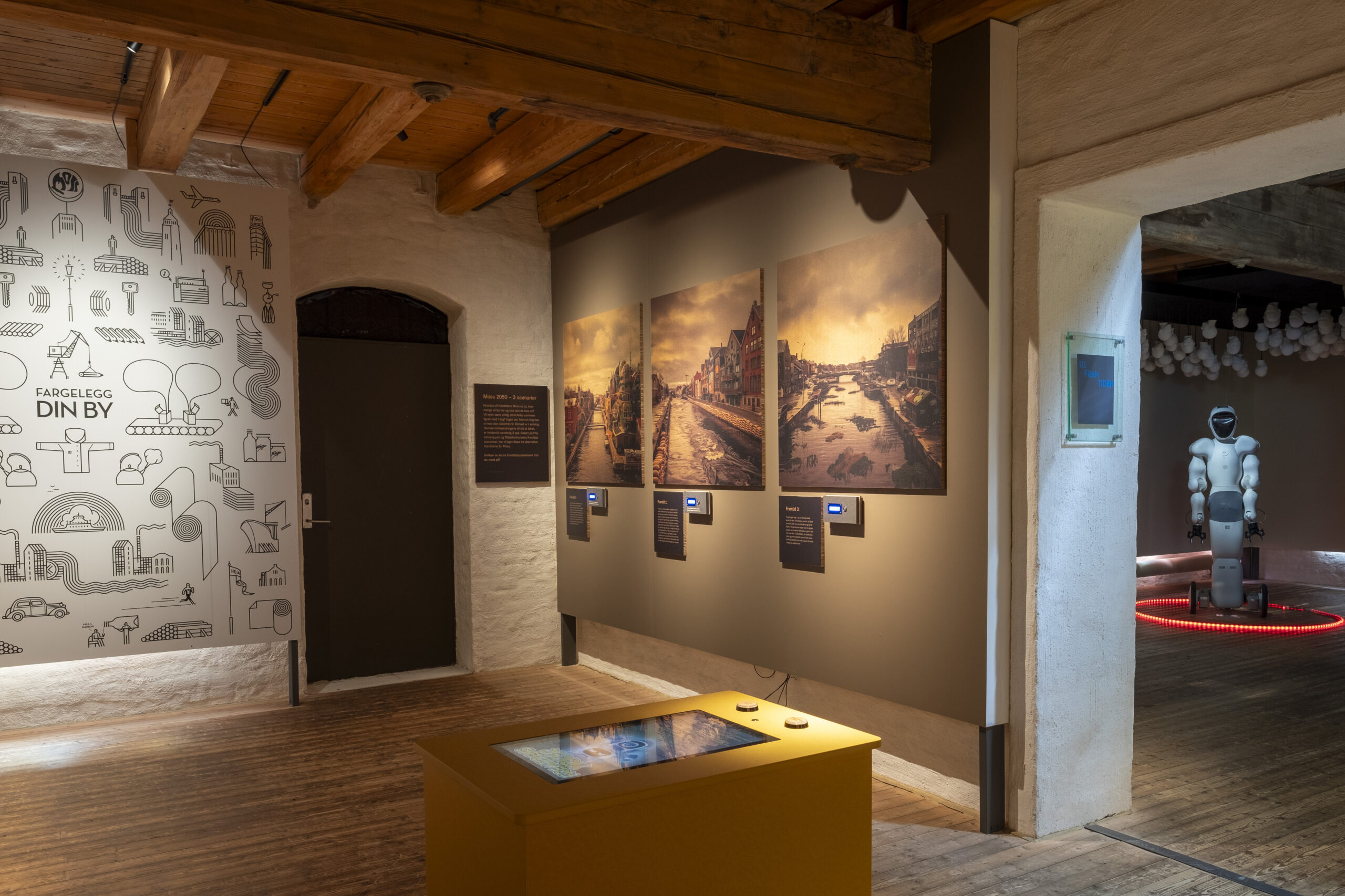 Exhibition space featuring the yellow console box for the interactive game 'Fremtidens Yrker,' surrounded by cityscape illustrations, photographic prints, and a humanoid robot in Moss Museum.